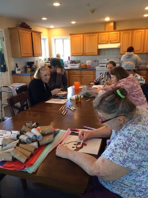 A group of elderly people sitting around a large wooden table in a kitchen, engaging in arts and crafts activities. One woman in the foreground is coloring a picture, while others are interacting or working on their own projects. The kitchen has wooden cabinets, a stove, and a window letting in natural light.