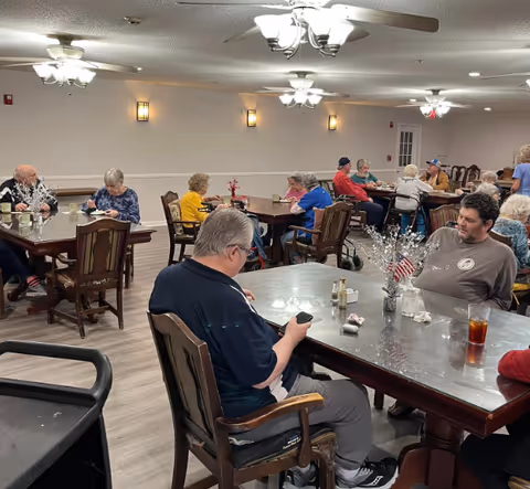 Older adults seated at multiple dining tables in a communal dining room with ceiling fans and simple table decorations.