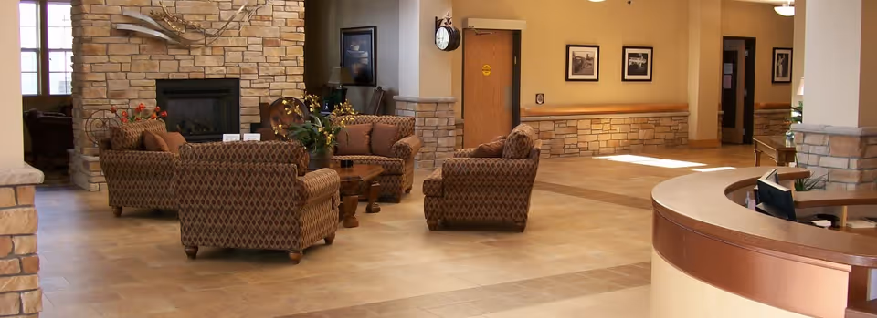 A spacious senior living facility common area with a stone fireplace, four patterned armchairs arranged around a wooden coffee table, and a curved reception desk in the foreground. The walls are decorated with framed pictures and the floor is tiled.