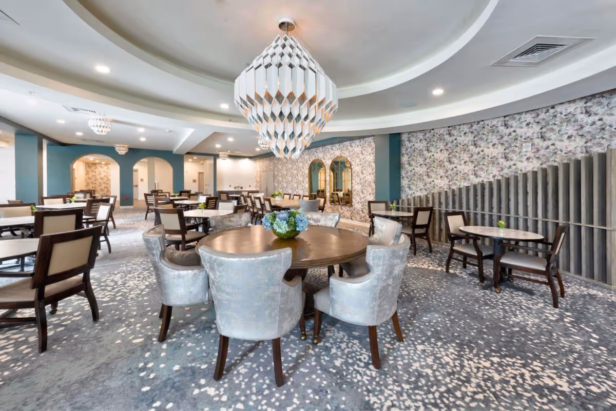 Bright upscale communal dining room with round tables, upholstered chairs, patterned carpet and a central chandelier.