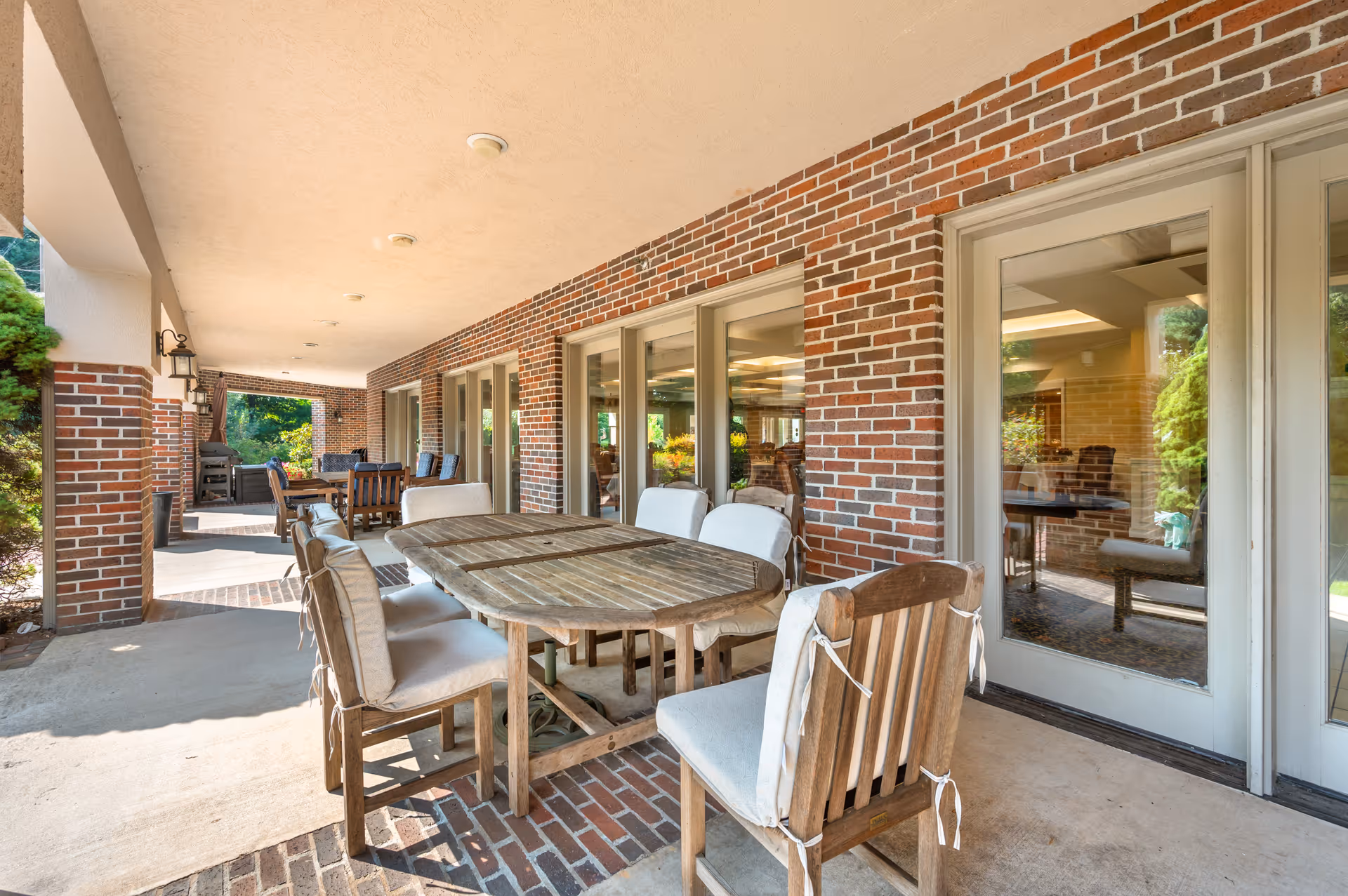 Covered outdoor patio area with wooden tables and cushioned chairs, brick walls, and large glass doors leading inside. Greenery is visible outside the patio.