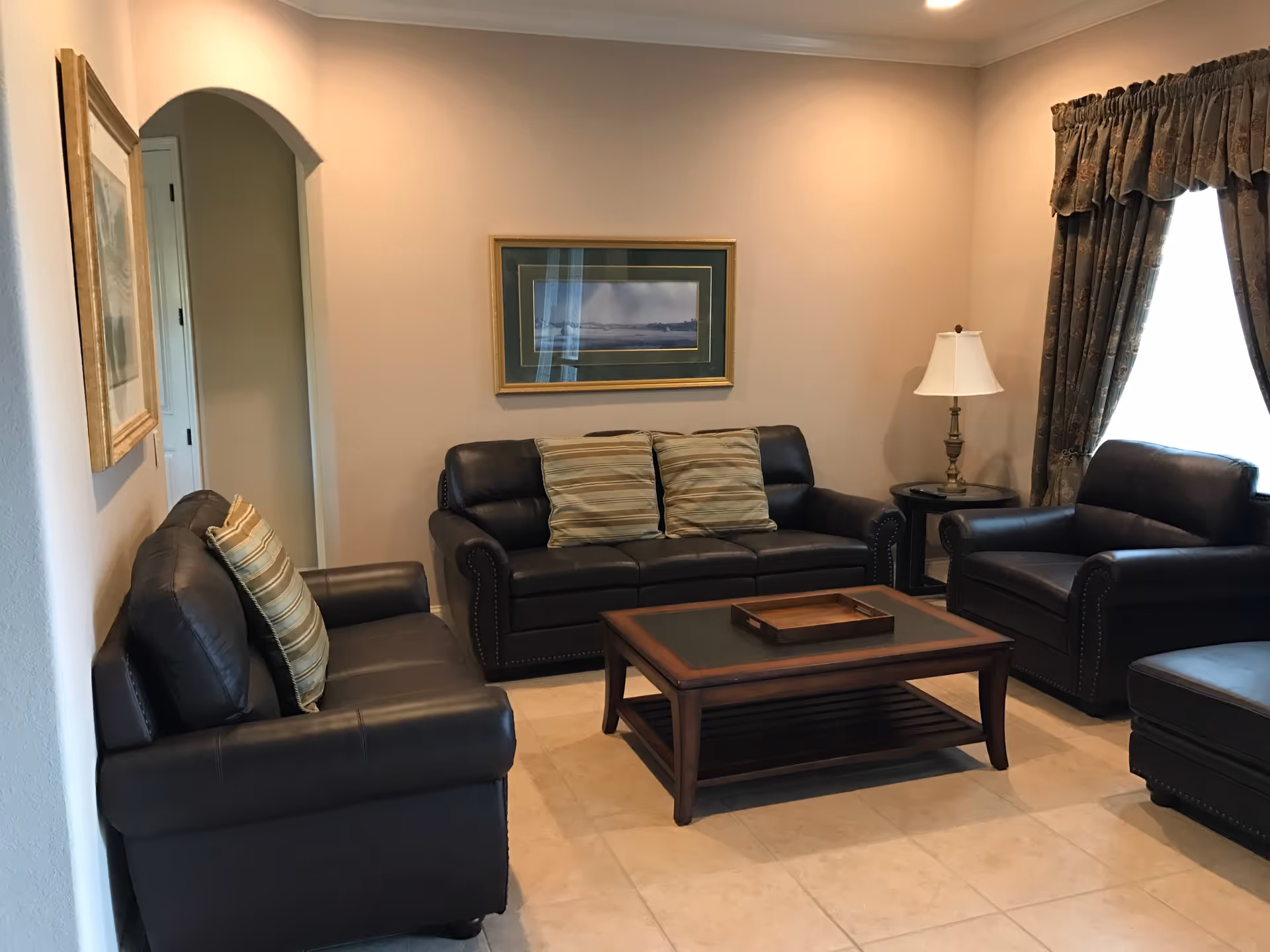 A cozy living room with three dark leather sofas arranged around a wooden coffee table with a tray on top. The room has beige tiled flooring, light-colored walls, a framed landscape painting on the wall, a side table with a lamp, and a window with patterned curtains.