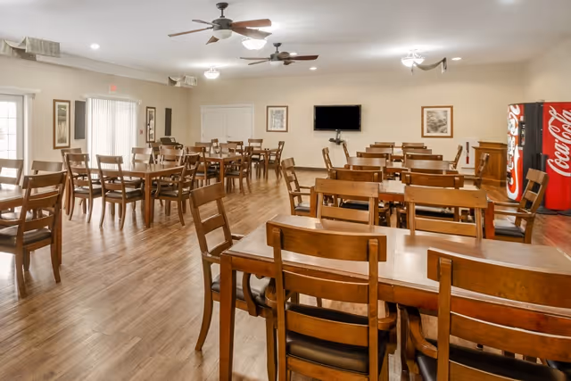 A spacious dining room with multiple wooden tables and chairs arranged neatly on a wooden floor. The room has ceiling fans with lights, framed artwork on the walls, a flat-screen TV mounted on the far wall, and a Coca-Cola vending machine in the corner near a wooden cabinet. Large windows with vertical blinds allow natural light to enter the room.