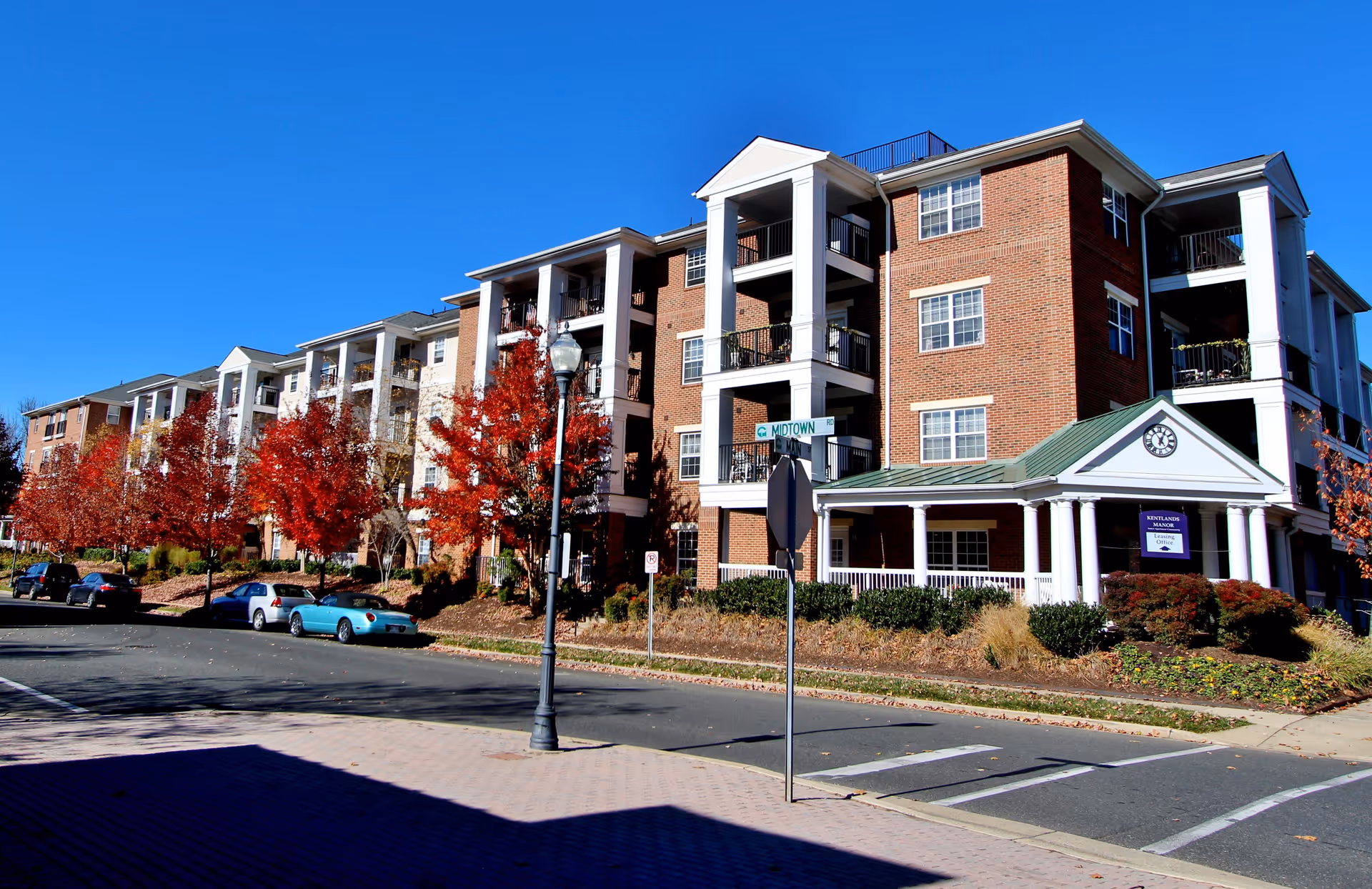 Exterior view of a multi-story brick residential building with white columns and balconies, surrounded by trees with red autumn leaves and parked cars along the street. A sign near the entrance reads 'Kentlands Manor Leasing Office.'