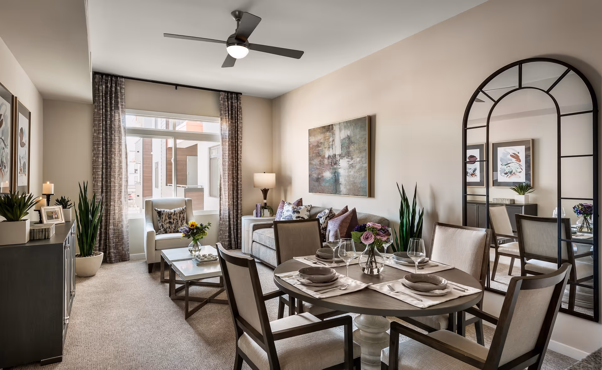 A modern living and dining area with a round dining table set for four, a gray sofa with decorative pillows, an armchair by a large window with curtains, a coffee table with a flower vase, indoor plants, wall art, and a large arched mirror reflecting part of the room.