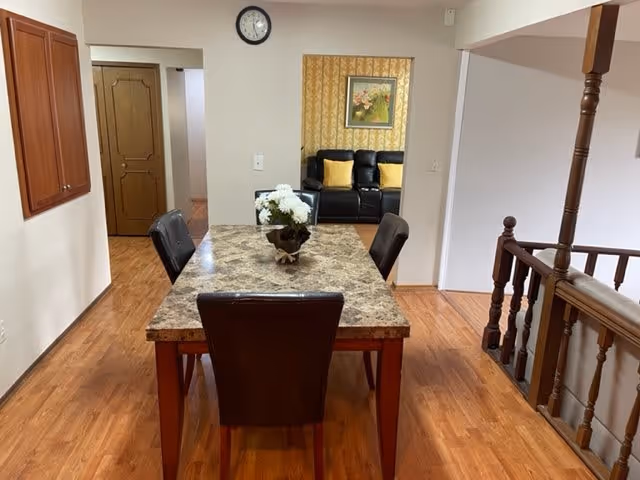 Interior view of a dining area with a rectangular marble-top table surrounded by four dark chairs. A vase with white flowers is placed on the table. The room has wooden flooring and light-colored walls. In the background, there is a doorway leading to a living room area with a black sofa and yellow cushions, a framed floral painting on the wall, and a clock above the doorway. To the right, there is a wooden railing with spindles and a staircase going down.