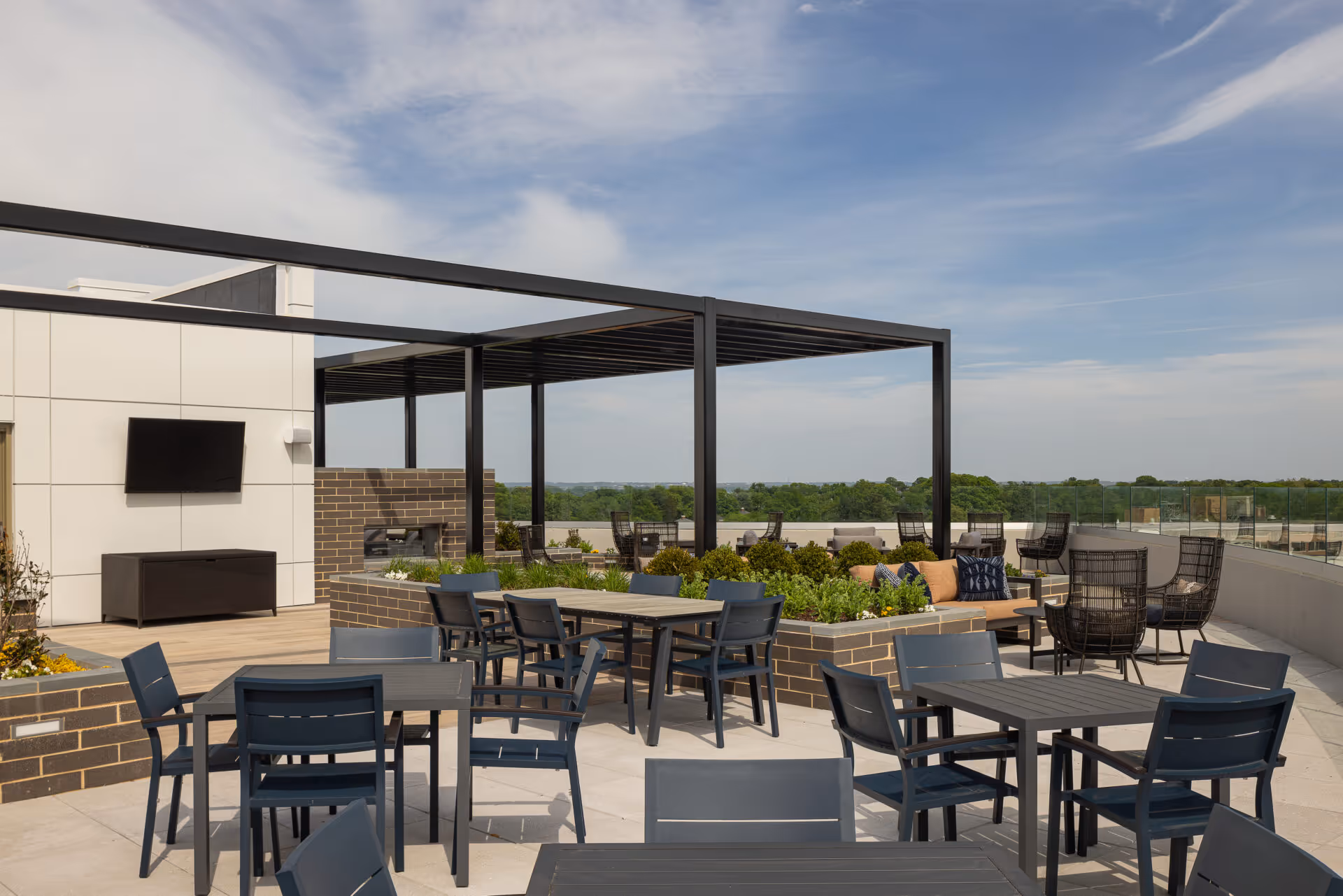 Outdoor patio area with multiple black tables and chairs, a pergola structure, brick planters with greenery, and a TV mounted on a white wall. The patio overlooks a scenic view with trees and a partly cloudy sky.