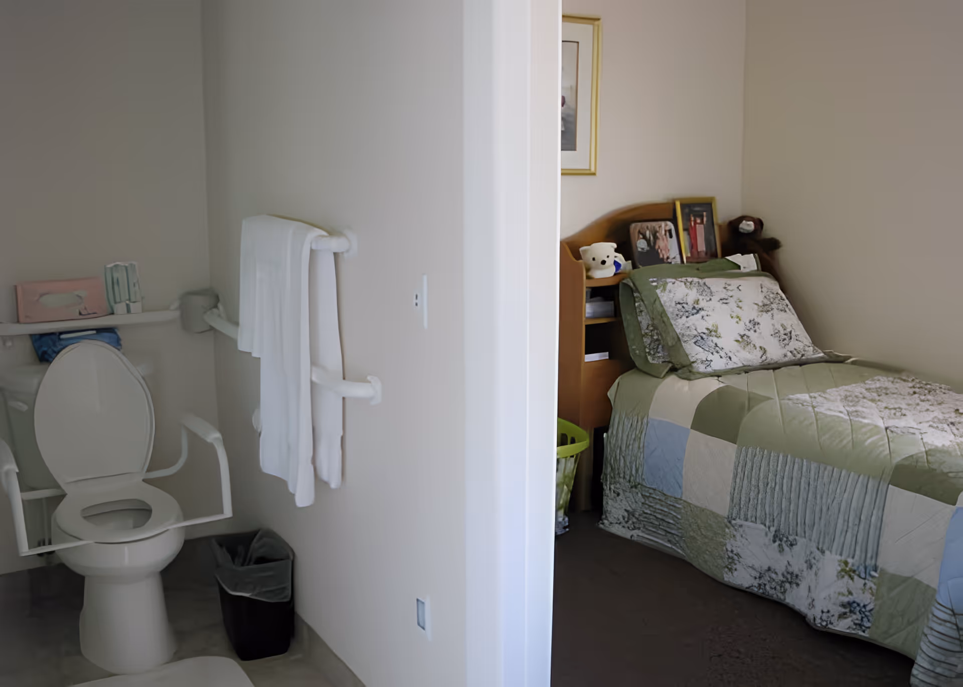 View of a small bedroom with a single bed covered in a green and blue patchwork quilt, adjacent to a bathroom with a toilet equipped with safety rails and a towel hanging on the wall.