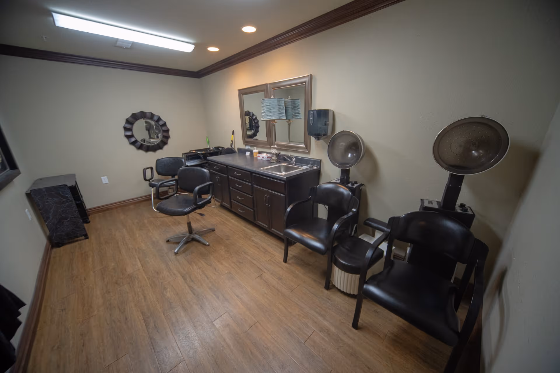 Interior view of a salon room with wooden flooring, two black salon chairs in front of a counter with a sink and two mirrors, and two black chairs with hair dryers attached on the right side.