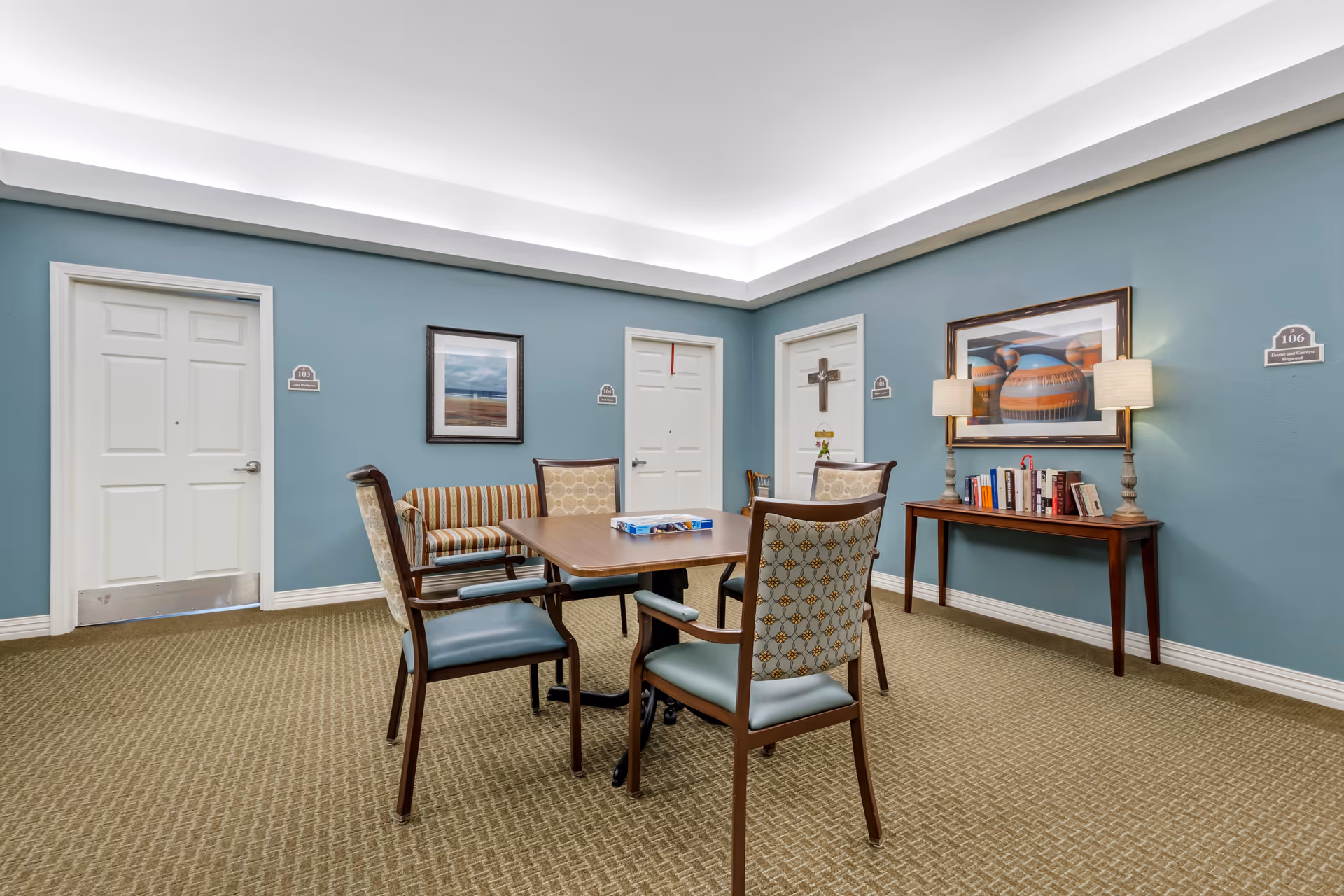A common area in a senior living facility with a square wooden table surrounded by four chairs with patterned upholstery. Behind the table, there is a striped loveseat against a blue wall with three white doors labeled 103, 104, and 105. A wooden console table with books and two lamps is placed against the right wall, which also has a framed picture of pottery. The carpet is beige with a textured pattern, and the ceiling has recessed lighting.