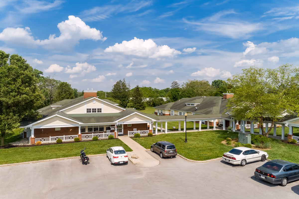 Front exterior of a single-story senior living facility with a covered walkway, parking lot, and landscaped lawns under a blue sky.