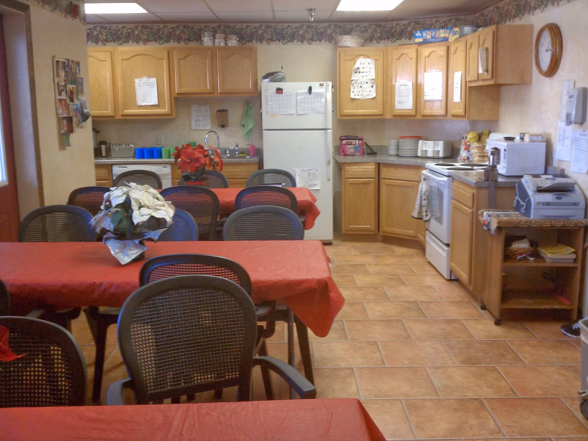 A communal kitchen and dining area with multiple tables covered in red tablecloths and black mesh chairs. The kitchen has wooden cabinets, a white refrigerator, a stove, a microwave, and various kitchen items on the counters. There are decorative plants on the tables and a clock on the wall.