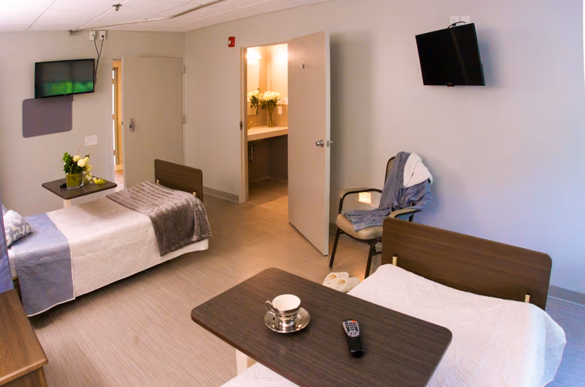 A room with two single beds, each with white and gray bedding. A small table with a cup and remote control is positioned between the beds. A chair with a robe draped over it is near the wall. Two wall-mounted televisions are visible, one on each side of the room. An open door reveals a bathroom with a sink and flowers on the counter.