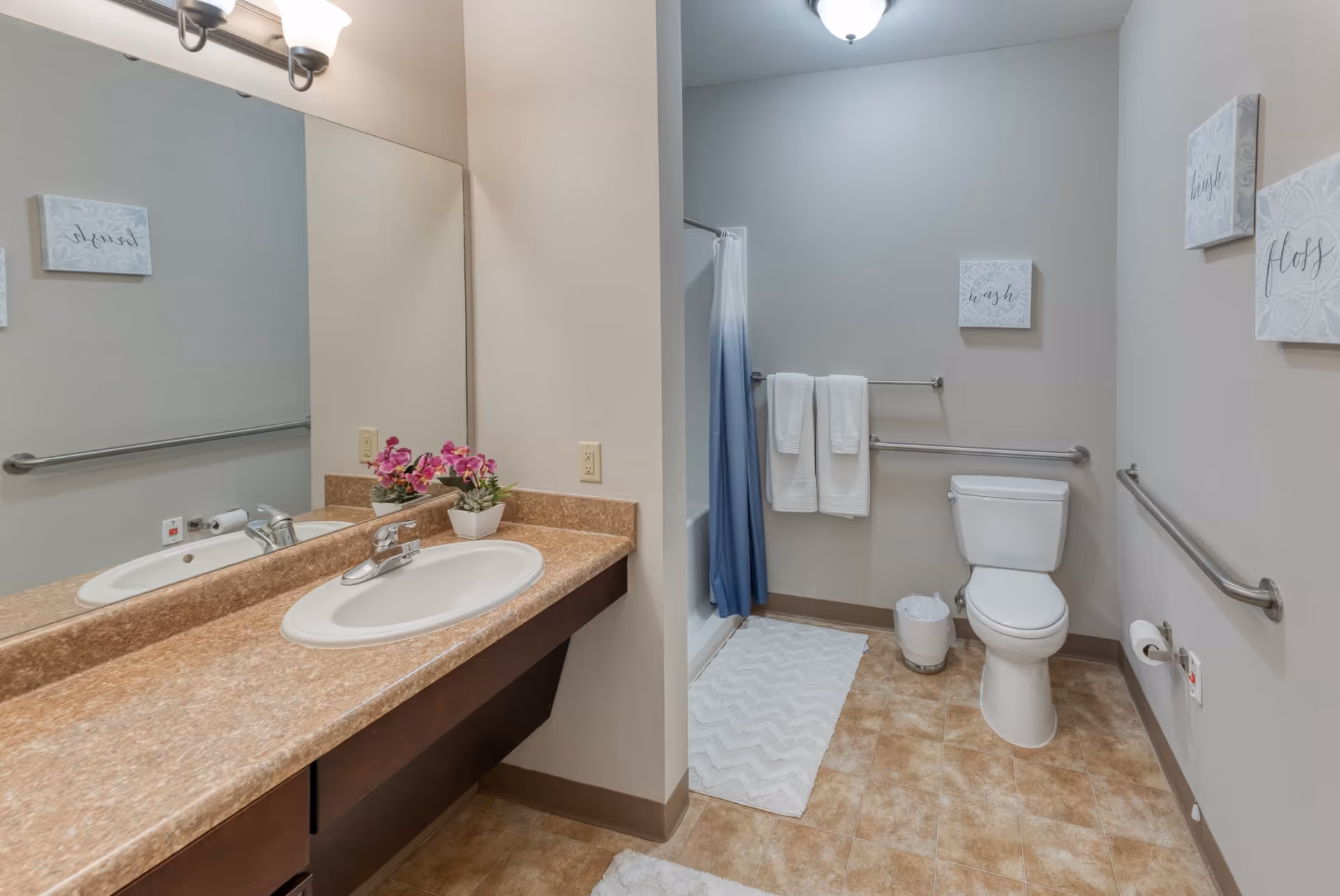 A clean and accessible bathroom with a single sink vanity featuring a brown countertop and a large mirror. There are two small potted plants with pink flowers on the counter. The bathroom has a toilet with grab bars on both sides and a shower with a blue and white gradient curtain. White towels hang on a towel rack above the toilet, and there are three decorative wall plaques with words like 'wash' and 'bless'. The floor is covered with beige tiles and white bath mats.