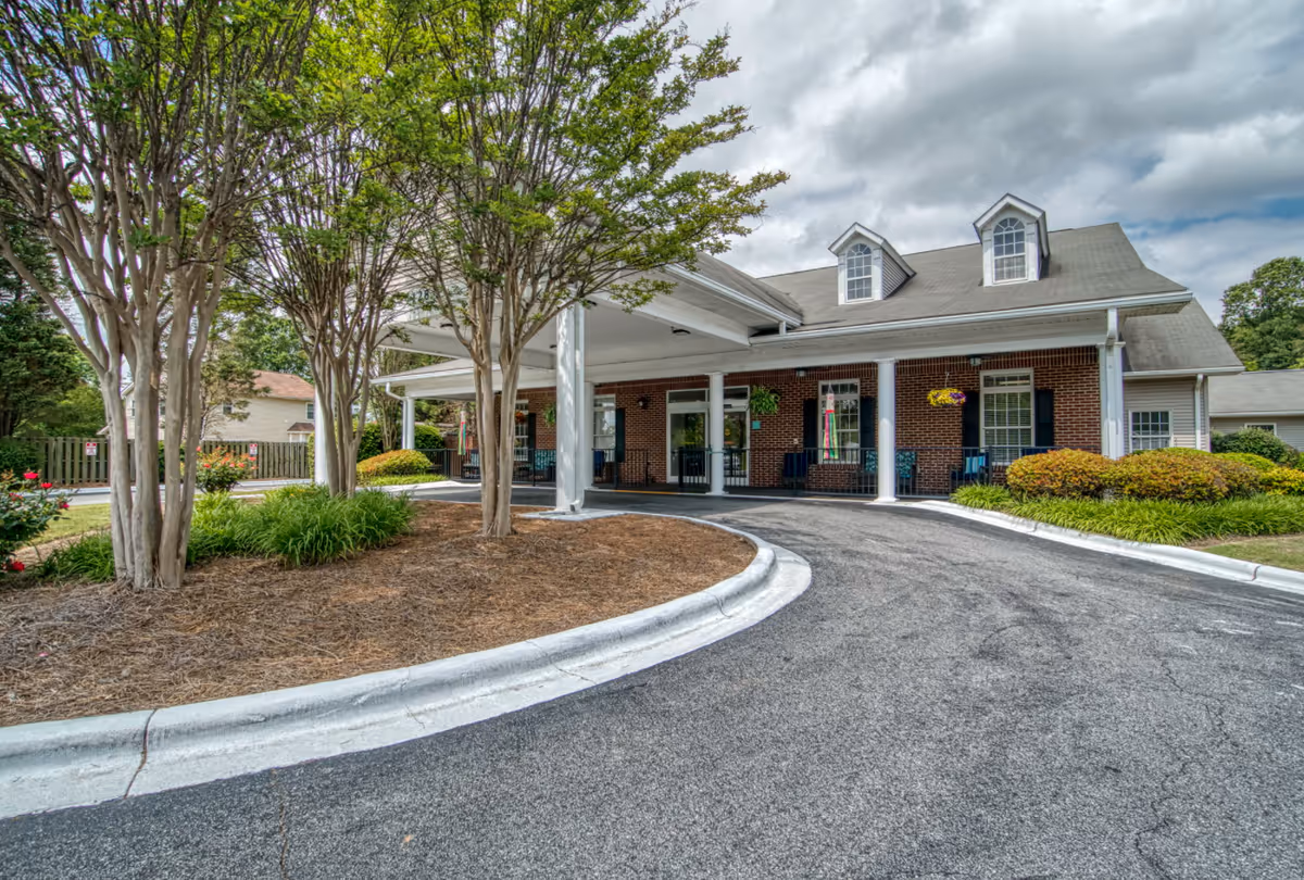 Exterior view of Summit Place of Mooresville showing a brick building with white columns and a covered entrance driveway. There are trees and landscaped bushes around the building under a cloudy sky.