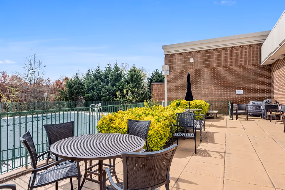 Outdoor patio area at The Charleston Senior Community with a round table and four chairs in the foreground, additional seating along the brick building wall, green bushes, and a fenced pool covered with a safety cover. Trees and a clear blue sky are visible in the background.