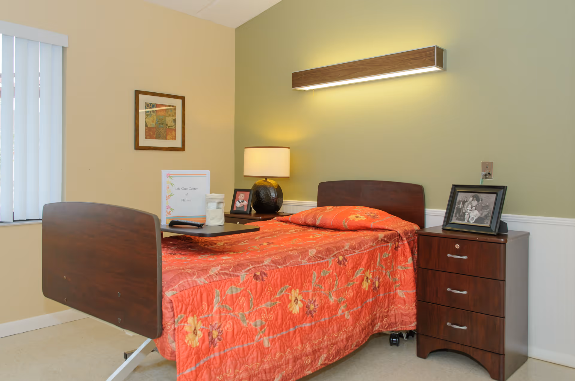 A senior care facility bedroom with a single bed covered in a red floral quilt, an overbed table, bedside lamp, nightstand with framed photos, and vertical blinds.