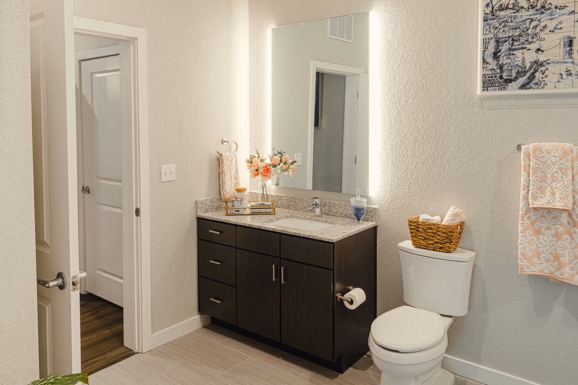 A modern bathroom with a dark wood vanity featuring a granite countertop and an illuminated rectangular mirror above it. On the countertop, there is a small tray with flowers and toiletries. To the right of the vanity is a white toilet with a wicker basket holding rolled towels on top. A peach and white patterned towel hangs on a wall-mounted towel rack. The bathroom has light-colored walls and floor tiles, and a door leading to another room is visible on the left.