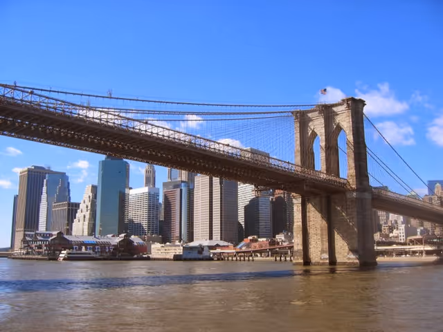 View of the Brooklyn Bridge spanning over the East River with the Manhattan skyline in the background under a clear blue sky.