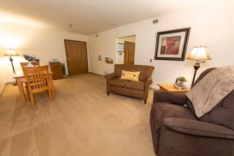 A cozy living area in a senior living facility featuring a brown patterned loveseat with a yellow pillow, a dark brown armchair with a gray throw blanket, a wooden side table with a lamp and plant, a wooden dining table with four chairs, and a framed artwork on the wall. The room has beige carpet and white walls.