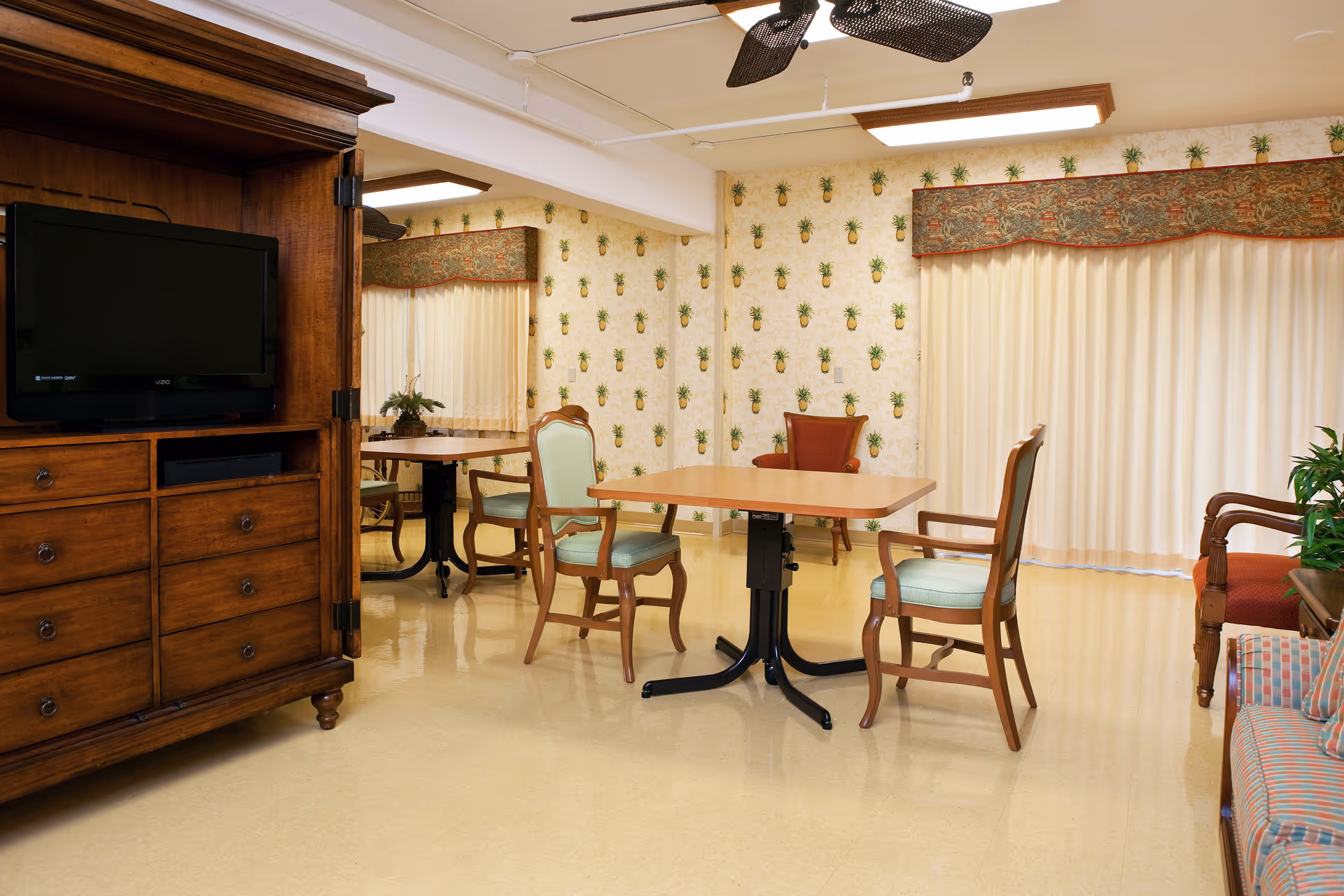 Interior common room with dining tables and chairs, a wooden TV armoire, patterned wallpaper, and large curtains.