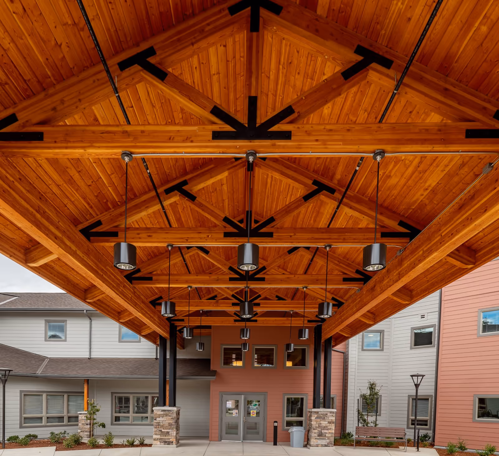 Entrance area of The Landing a Senior Living Community featuring a large wooden canopy with exposed beams and hanging lights. The building exterior has a combination of light gray and pink siding with multiple windows and a double door entrance.