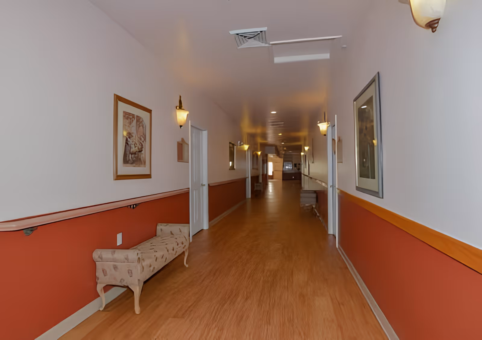Long, well-lit hallway in a retirement home with wooden floors, wall sconces, framed art, and a bench along the wall.