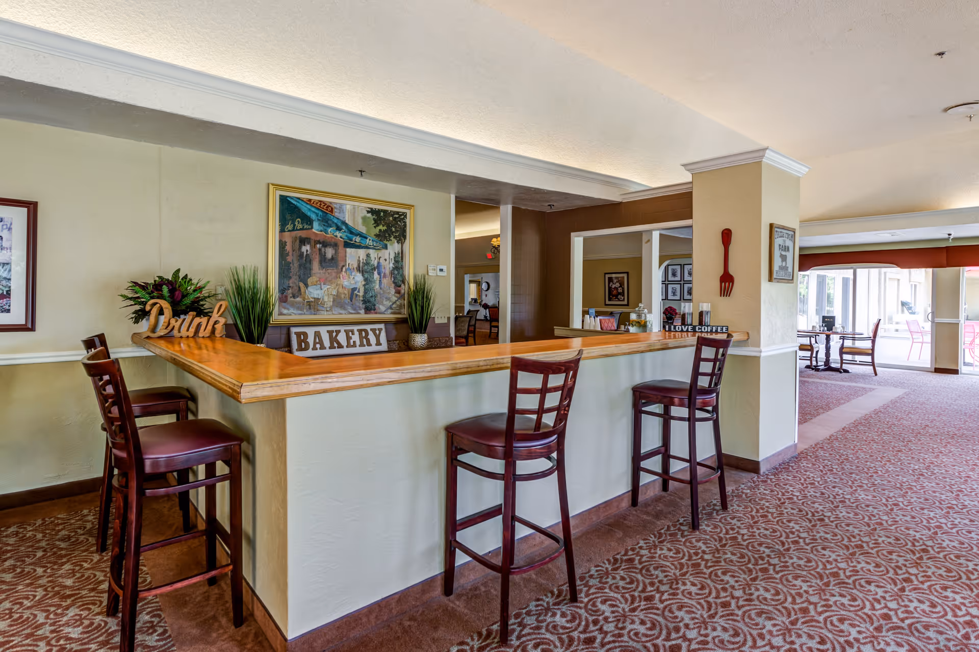 Interior view of a senior living facility lounge area with a wooden bar counter and four high chairs. The bar counter has decorative signs that say 'Drink' and 'Bakery', along with potted plants. The room has patterned carpet, light-colored walls, and framed artwork. In the background, there is a seating area with tables and chairs near large windows letting in natural light.