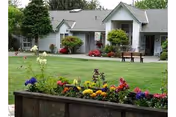 A well-maintained outdoor garden area in front of a single-story residential building with a gray roof and white walls. The garden features colorful flowers in a raised flower bed, green grass, shrubs, and a wooden bench near the building entrance.