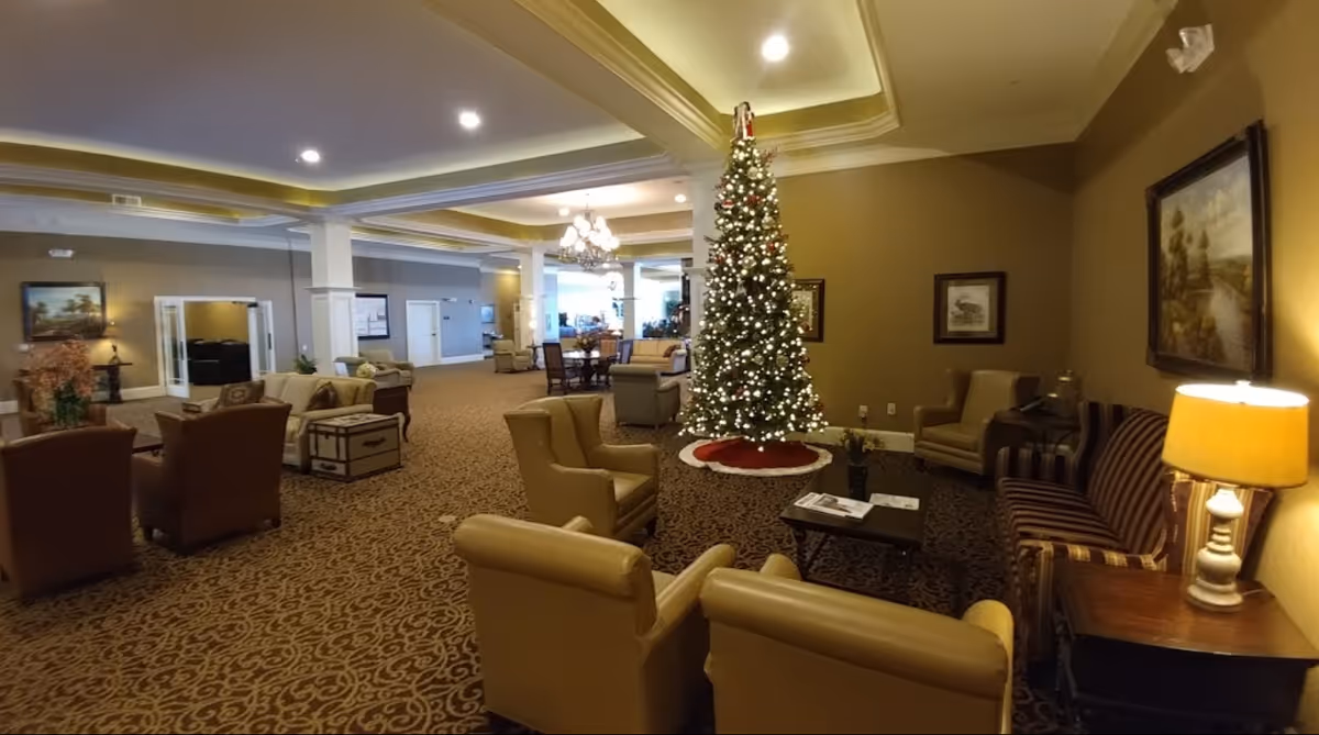 A spacious and warmly lit living room area decorated for Christmas with a tall, lit Christmas tree in the center. The room features multiple comfortable armchairs and sofas arranged around coffee tables, with framed artwork on the walls and a patterned carpet. There are ceiling lights and a chandelier providing illumination.