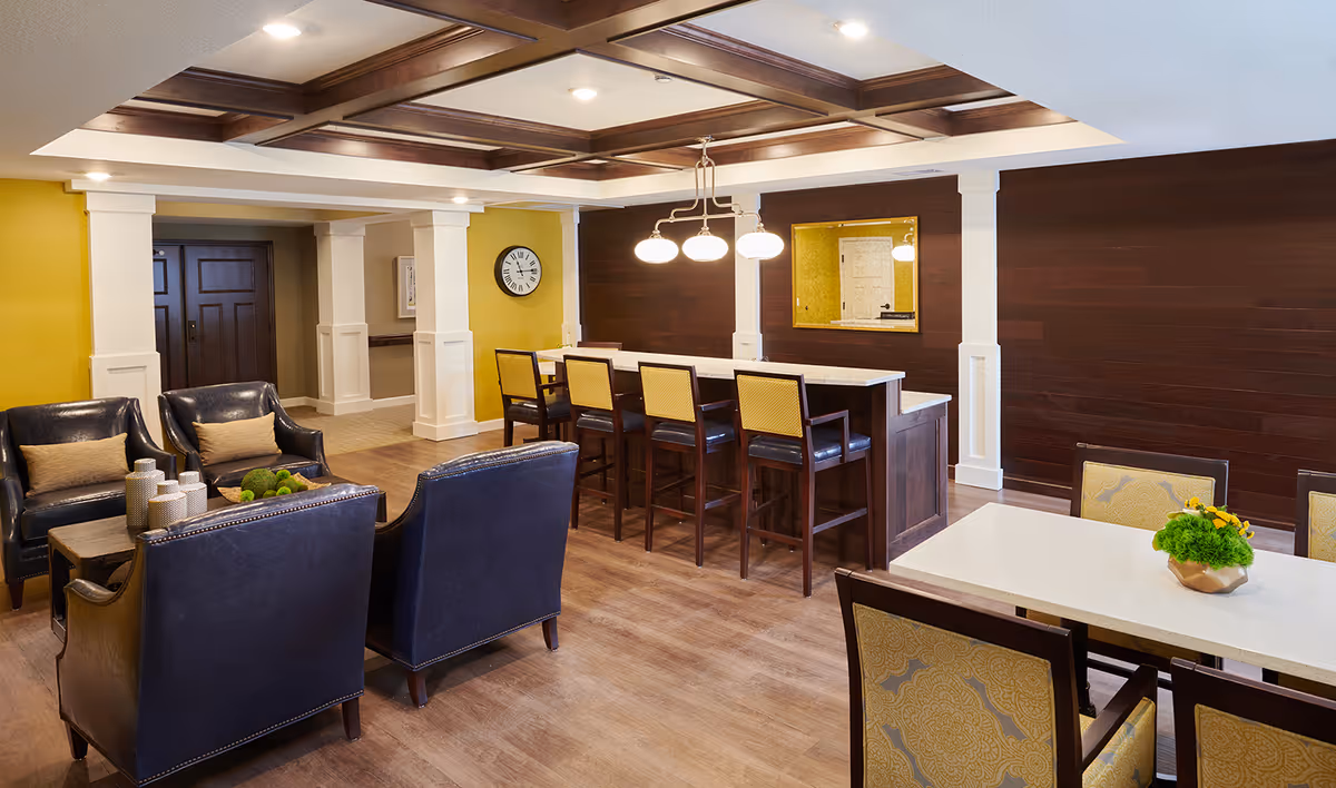 A cozy common area with dark leather armchairs and a wooden coffee table with decorative items. There is a high counter with four bar stools, a dining table with chairs, and a yellow accent wall with a clock. The ceiling has wooden beams with recessed lighting.