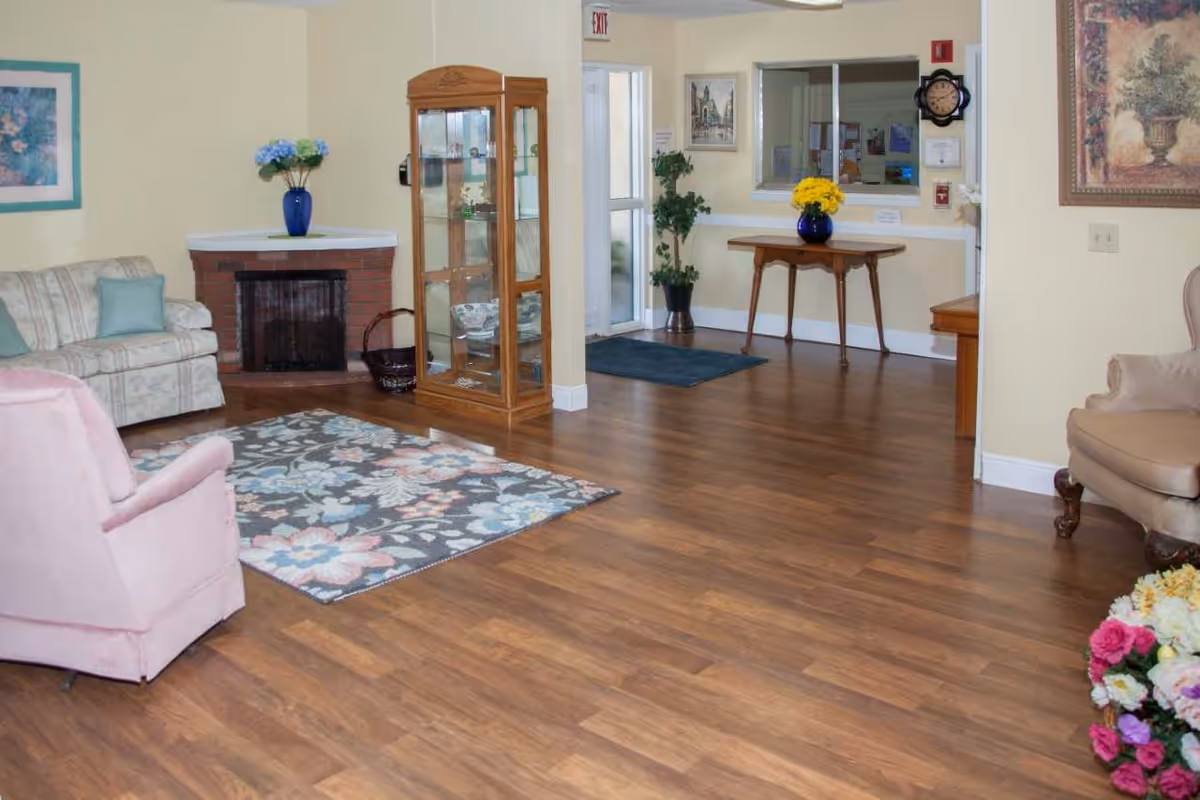 A bright common living room with seating, a floral rug, wooden floors, a display cabinet, and a table with flowers near a reception window.