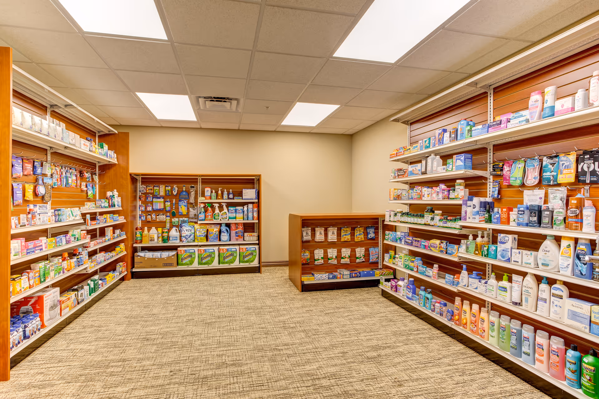 Interior view of a small retail shop area within a facility, featuring shelves stocked with various personal care products, toiletries, and household items. The room has beige walls, a carpeted floor, and a drop ceiling with fluorescent lighting.