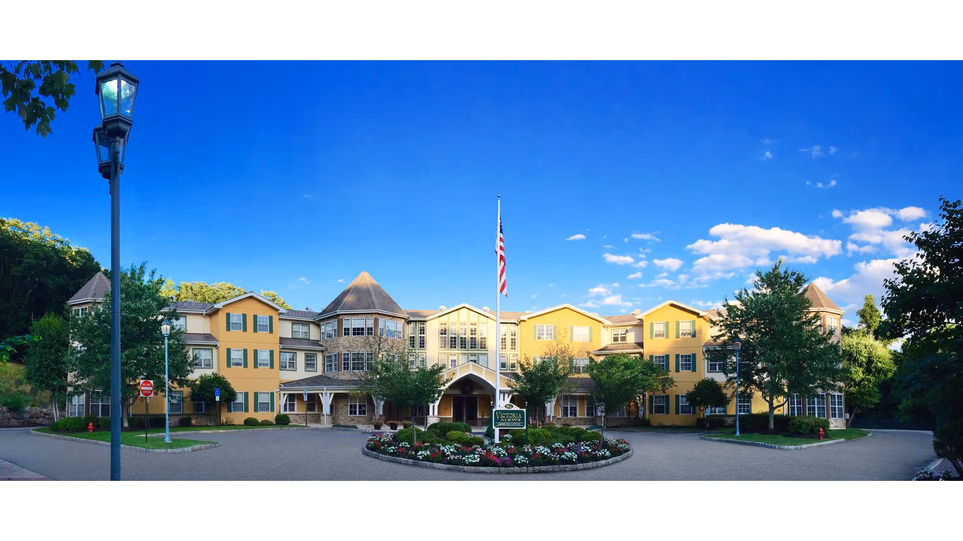 Wide exterior view of Victoria Mews Assisted Living facility, a large yellow three-story building with green shutters, surrounded by trees and landscaping. There is a circular driveway with a flower bed and an American flagpole in front of the main entrance under a covered porch. The sky is clear and blue with some scattered clouds.