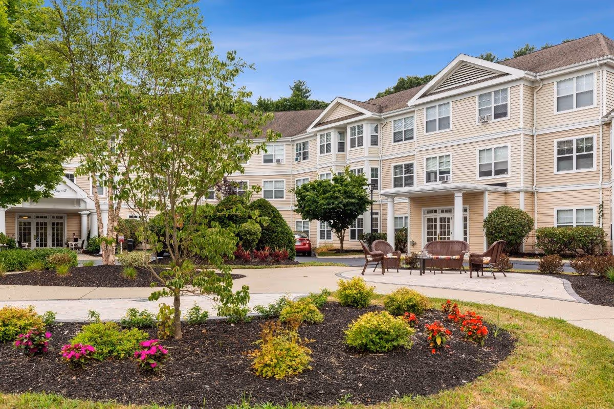 Exterior view of Benchmark Senior Living at Forge Hill showing a three-story beige building with white trim, surrounded by landscaped gardens with small trees, bushes, and colorful flowers. There is a paved walkway and outdoor seating area with chairs and a table in front of the building under a clear blue sky.