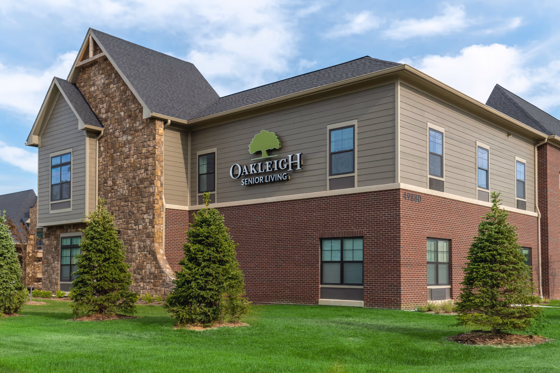 Exterior view of Oakleigh Senior Living building featuring a combination of brick, stone, and siding with multiple windows and a green lawn with small evergreen trees in front.