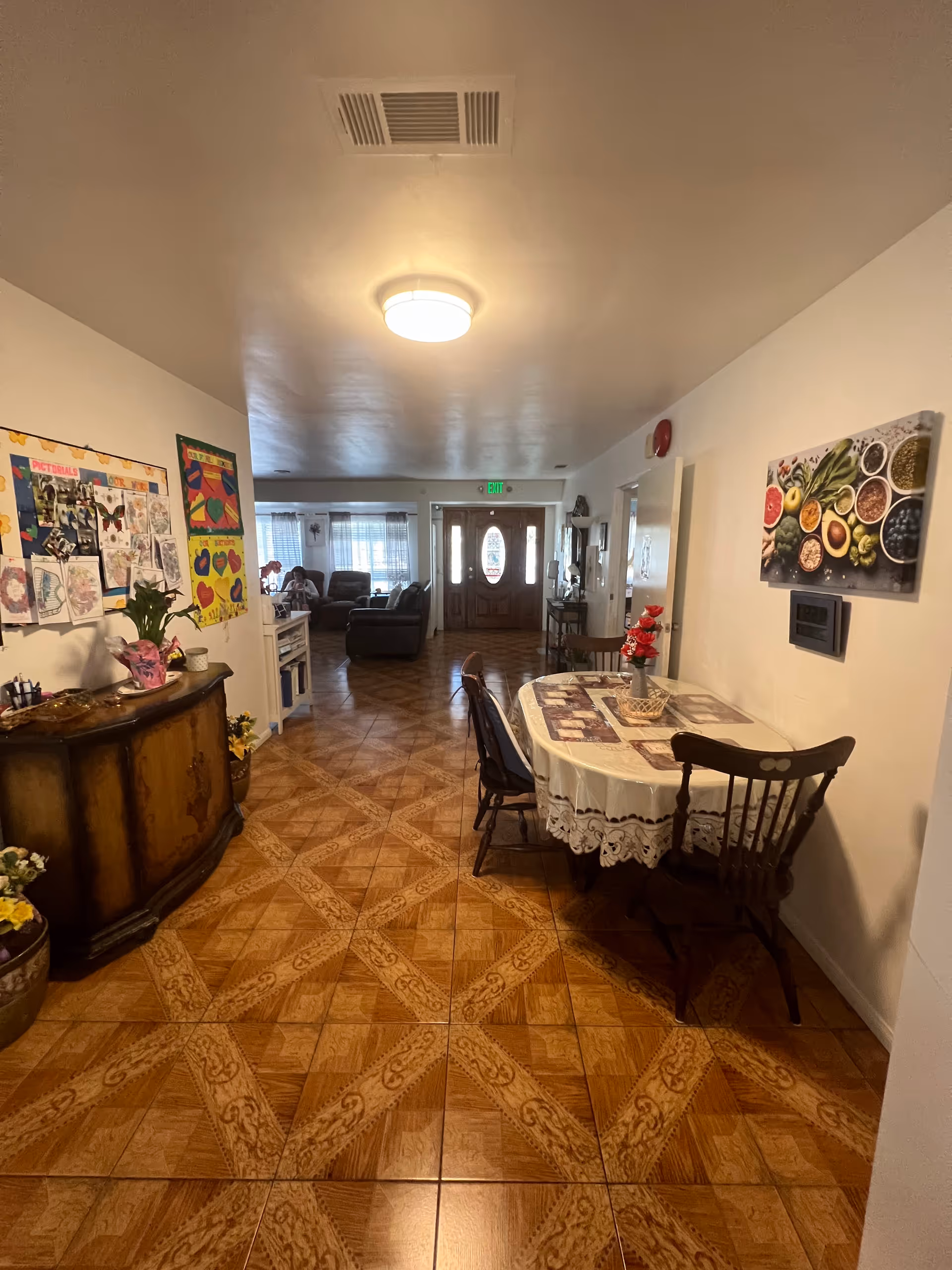 Interior view of a senior living facility hallway with a wooden table covered with a lace tablecloth and chairs on the right. On the left side, there is a wooden cabinet with flowers and bulletin boards on the wall. At the end of the hallway, there is a living room area with sofas and a front door with glass panels.