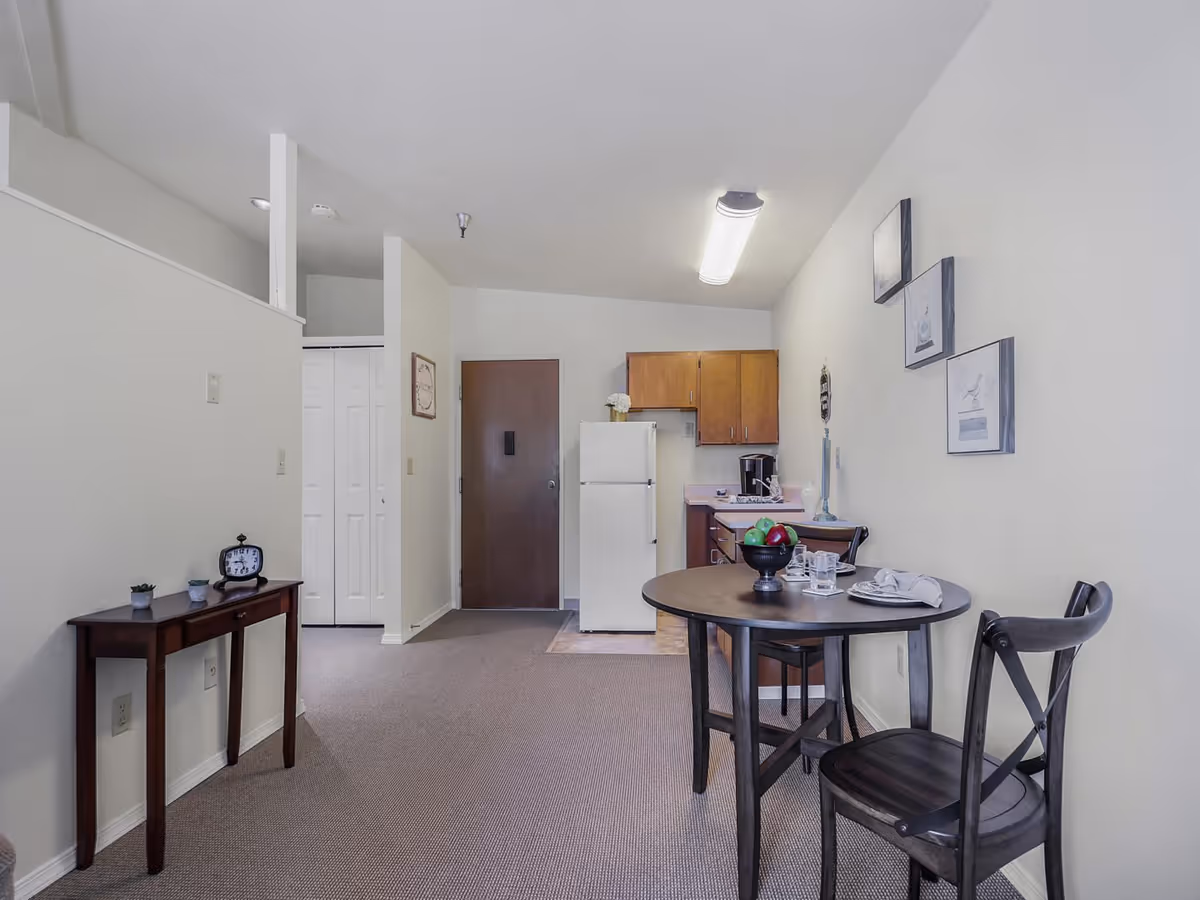 Small studio apartment interior with a round dining table and chairs, a kitchenette with refrigerator and cabinets, and an entry door.