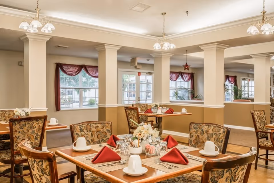 Dining area with multiple tables set with white cups, glasses, silverware, and red folded napkins. The chairs have floral upholstery. The room has beige walls with white trim, large windows with red curtains, and hanging light fixtures.