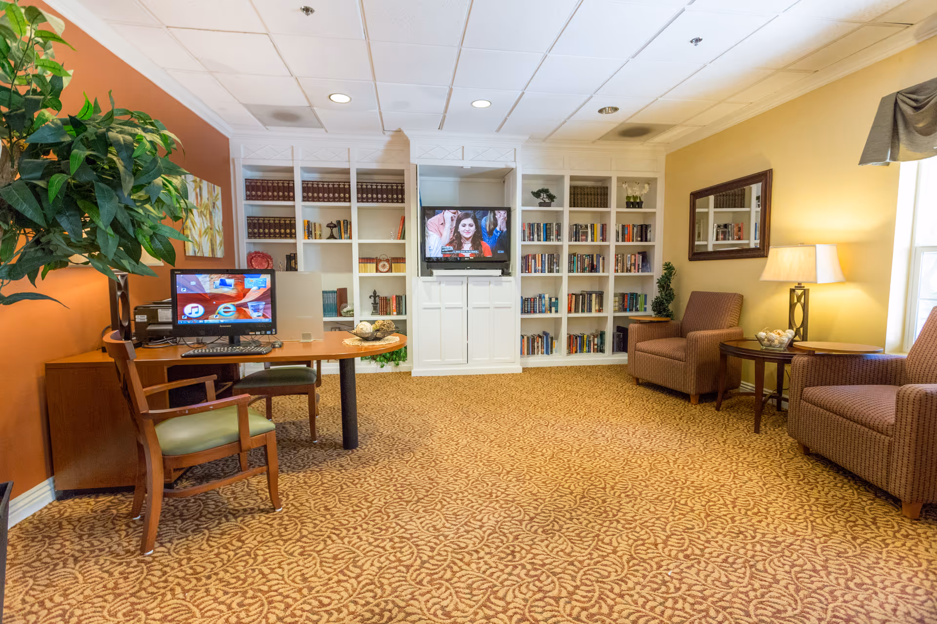 A cozy living room area in a senior living facility featuring a patterned carpet, two upholstered armchairs with a small round table and lamp between them, a large built-in white bookshelf filled with books and decorative items, and a flat-screen TV mounted in the center of the bookshelf. On the left side, there is a wooden desk with a computer and chair, a large green plant, and a warm orange accent wall with artwork.