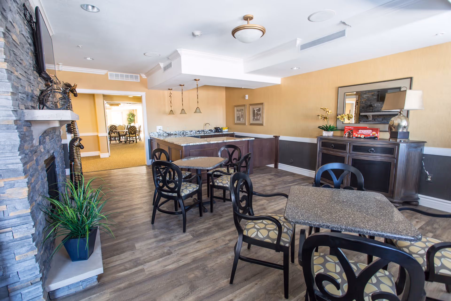 Bright communal dining/lounge area with multiple tables and chairs, a stone fireplace, and a serving counter.