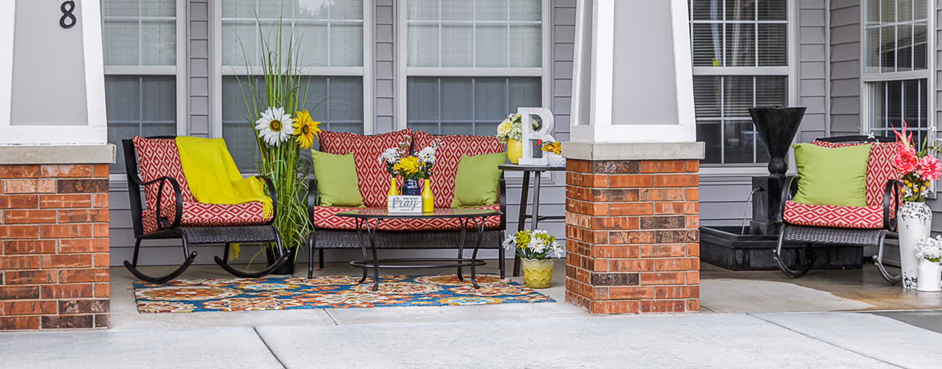 Outdoor seating area with red patterned cushioned rocking chairs and a matching cushioned bench with green pillows and a yellow throw blanket. The seating is arranged on a colorful rug between brick pillars in front of a building with large windows. There are decorative flowers and a small table with a vase and letter 'P'.