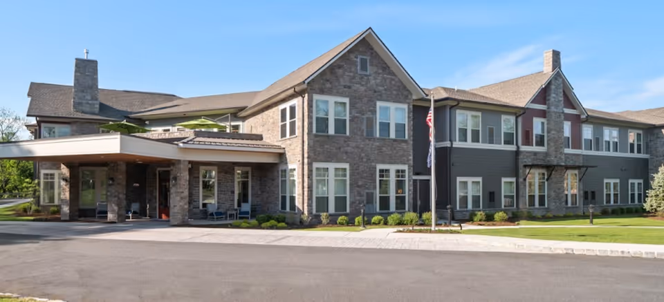 Exterior view of a two-story assisted living facility building with stone and siding facade, multiple windows, a covered entrance, and a flagpole with an American flag in front. The building is surrounded by a paved driveway and well-maintained landscaping under a clear blue sky.