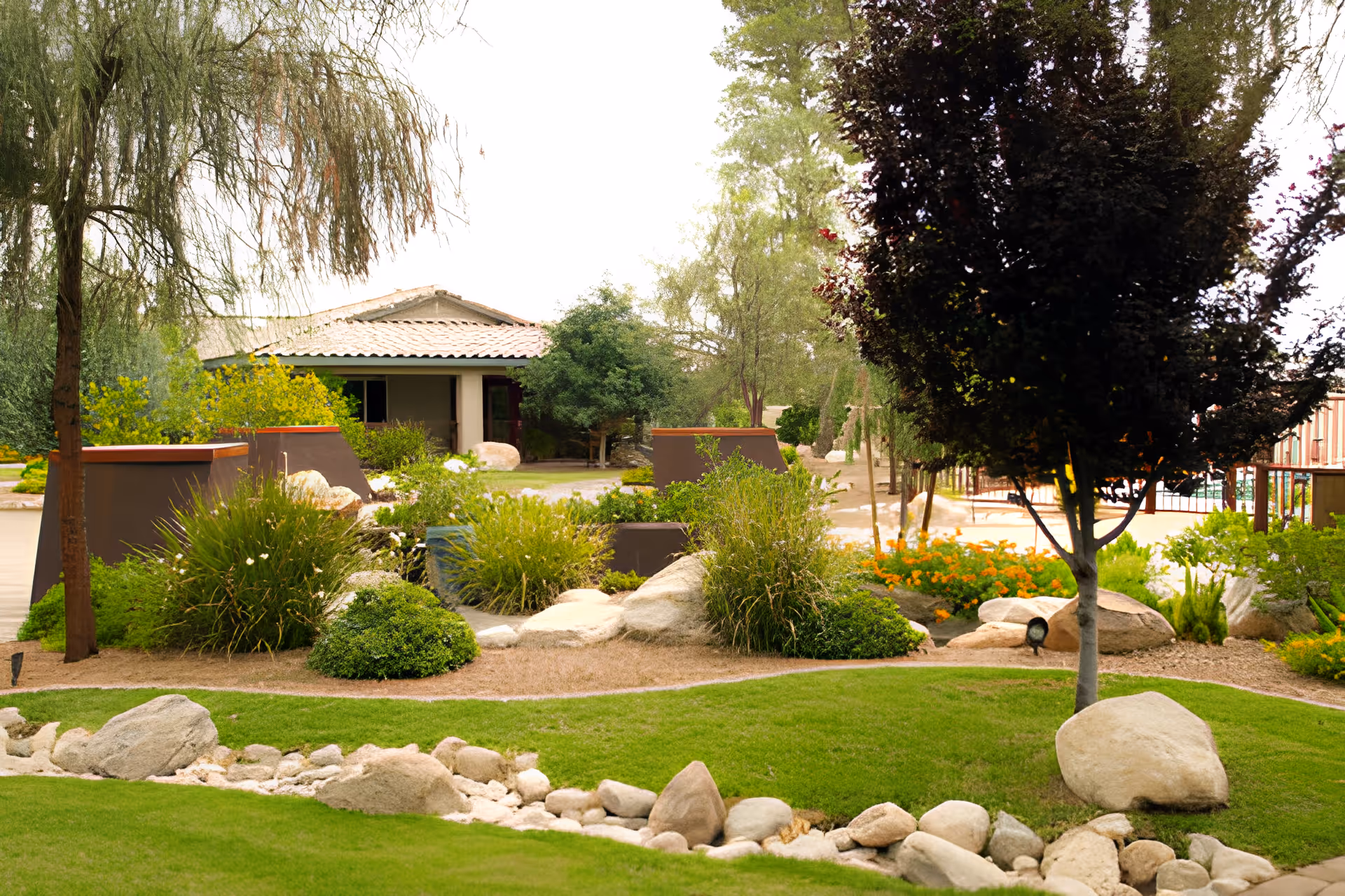 A landscaped outdoor garden area with green grass, various bushes, rocks, and trees. In the background, there is a building with a tiled roof partially visible behind the greenery.