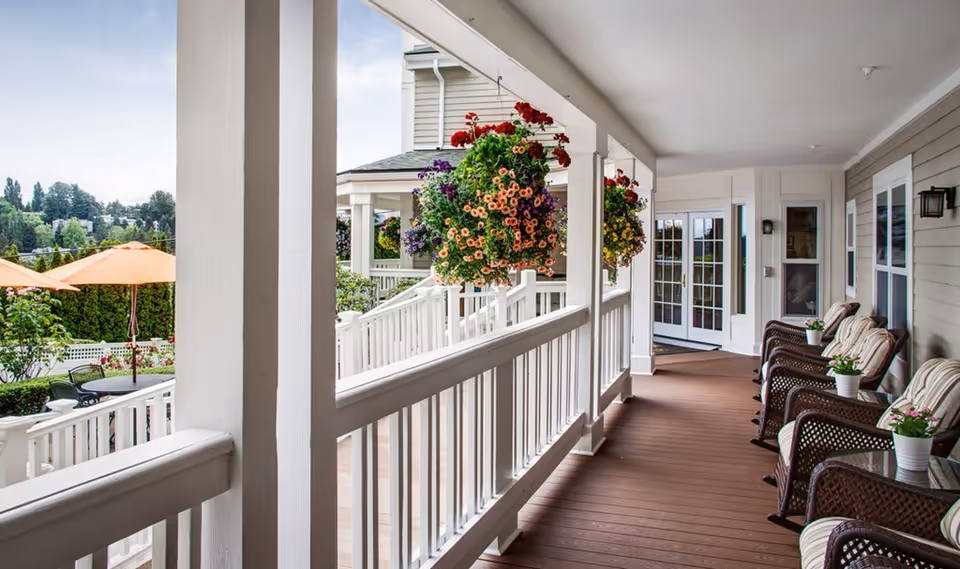 Covered porch area with wicker chairs and small tables, each table holding a potted plant. Hanging flower baskets with colorful blooms are suspended from the porch ceiling. In the background, there are orange patio umbrellas and greenery, suggesting a garden or outdoor seating area.