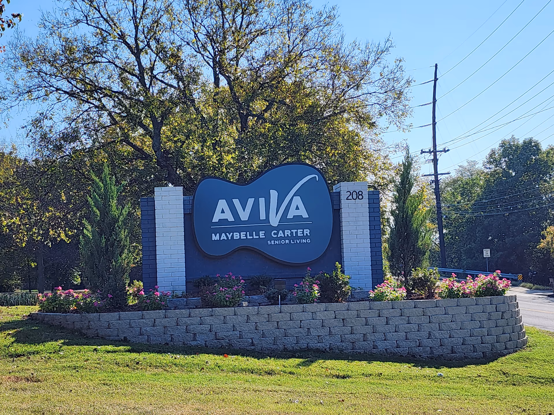 Outdoor view of a large sign for AVIVA Maybelle Carter Senior Living, surrounded by a landscaped area with flowers, small trees, and a stone retaining wall. Trees and power lines are visible in the background under a clear blue sky.