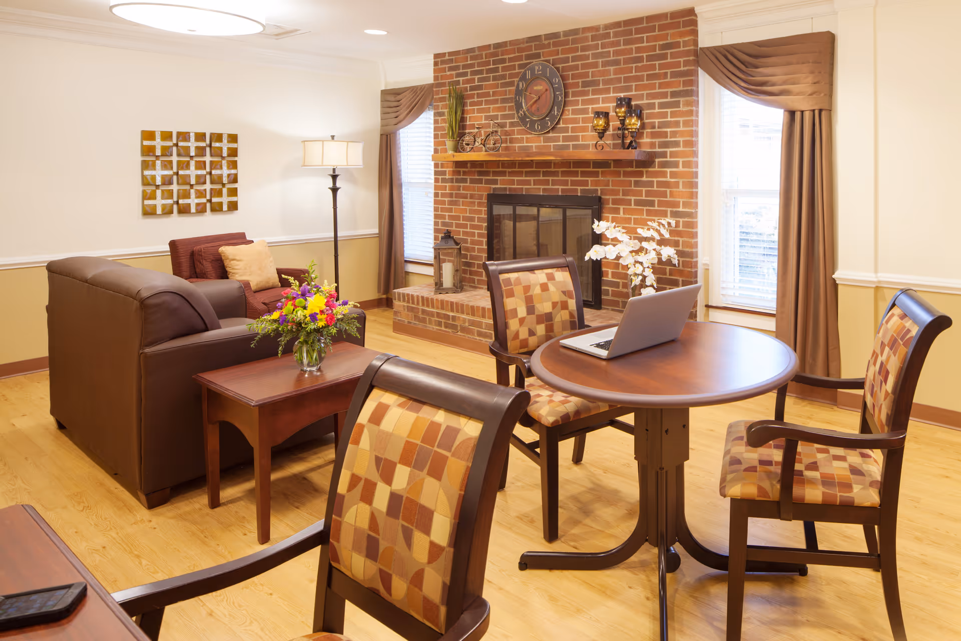 A cozy living room with a brick fireplace, a round wooden table with three patterned chairs, a laptop on the table, a brown leather sofa, an armchair with a cushion, a side table with a vase of flowers, a floor lamp, and two windows with brown curtains.