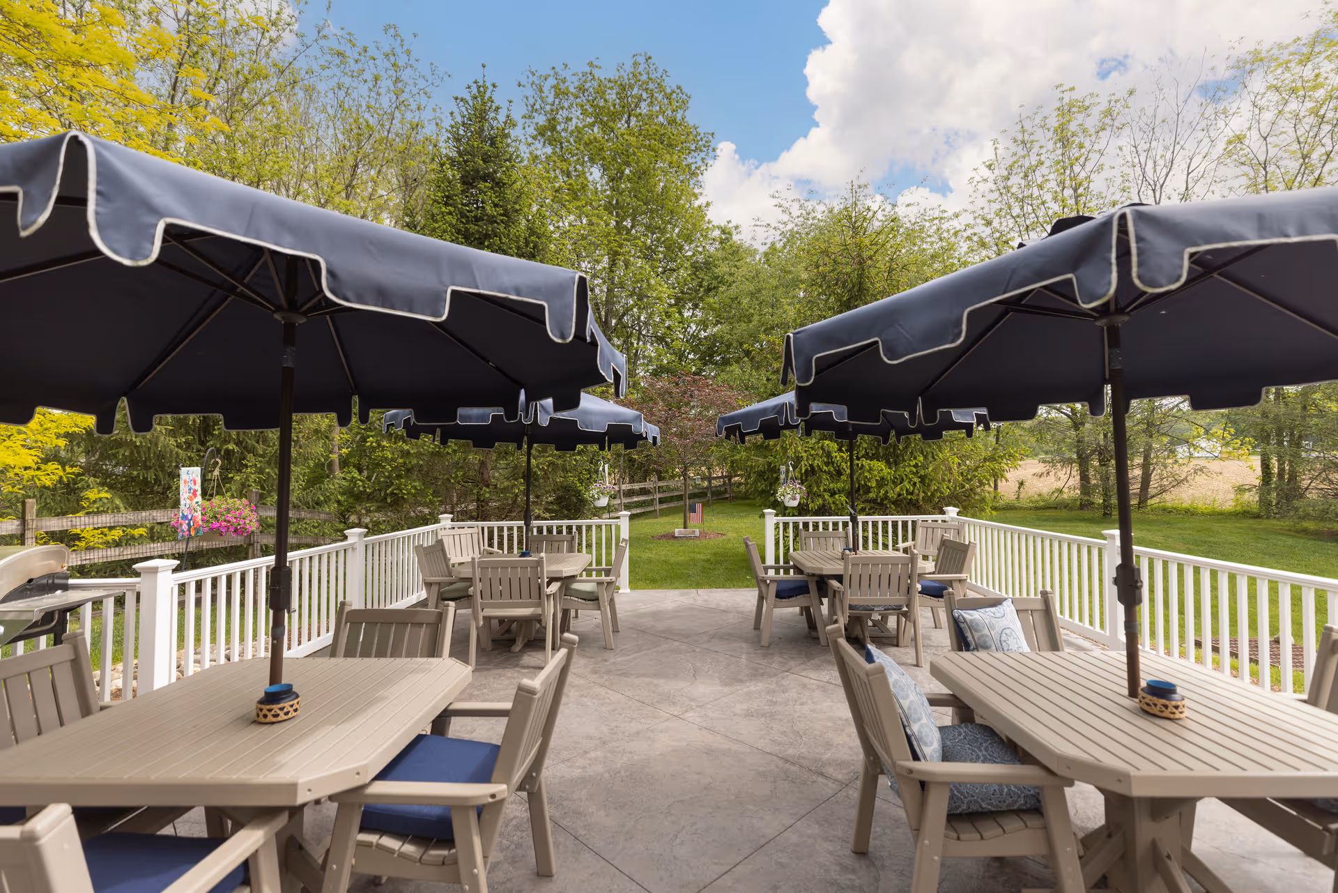 Outdoor patio area with several beige tables and chairs, each table shaded by large dark blue umbrellas. The patio is surrounded by a white railing and overlooks a green lawn with trees and a partly cloudy sky.