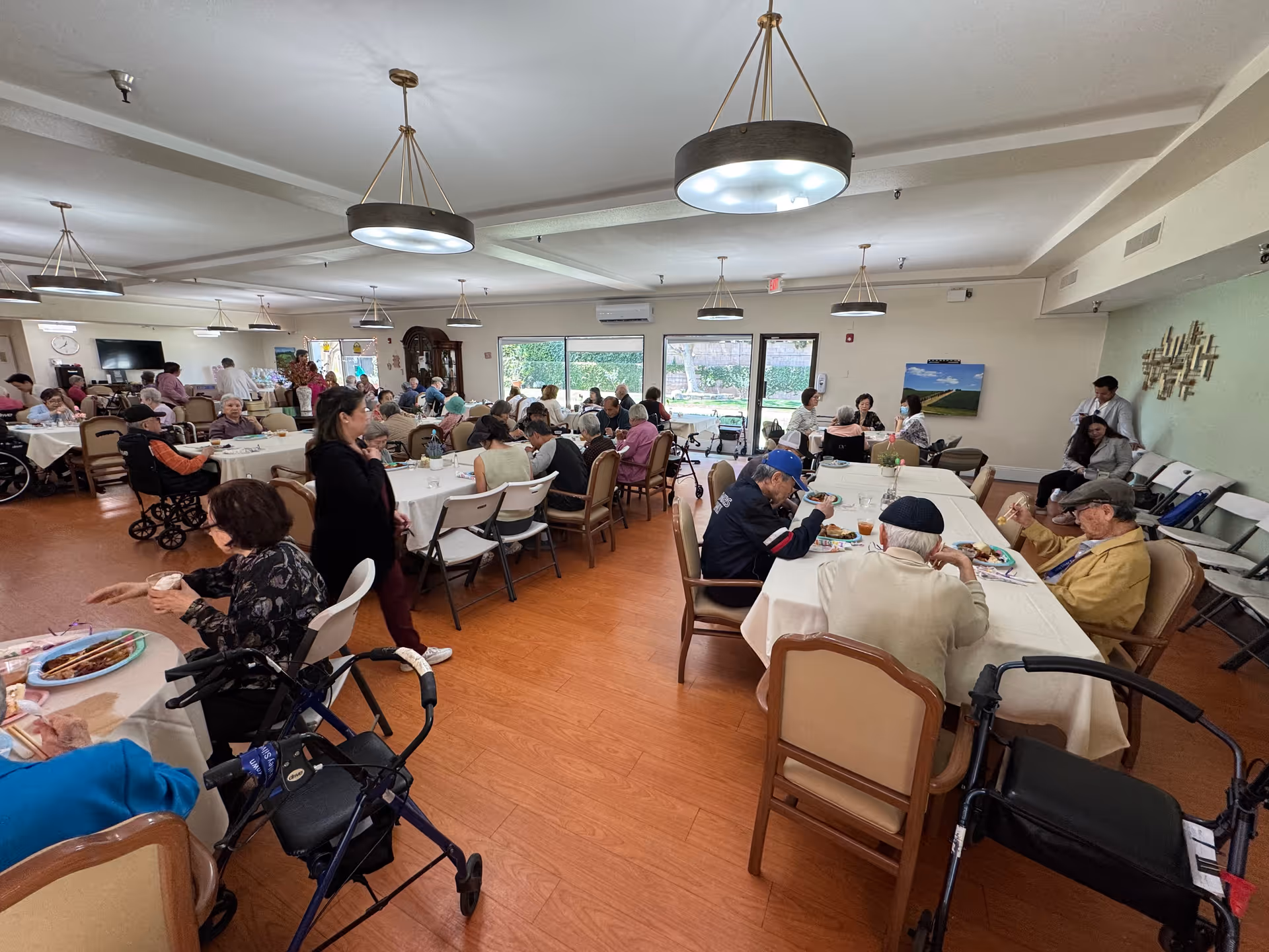 A large dining room filled with elderly residents seated at tables covered with white tablecloths, eating and socializing. Several walkers and wheelchairs are visible. The room has wooden flooring, large windows letting in natural light, and modern ceiling lights. Staff members are also present assisting residents.