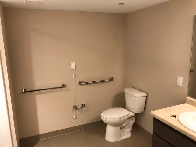 A clean, accessible bathroom with beige walls and floor. The image shows a white toilet with grab bars on the adjacent walls for support. There is a pull cord emergency alarm on the wall next to the toilet. To the right, part of a countertop with a sink and a mirror above it is visible.
